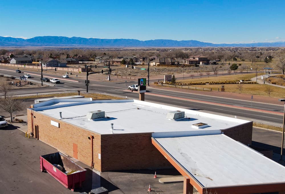 Aerial view of a commercial building with a flat roof, surrounded by residential areas and mountains.